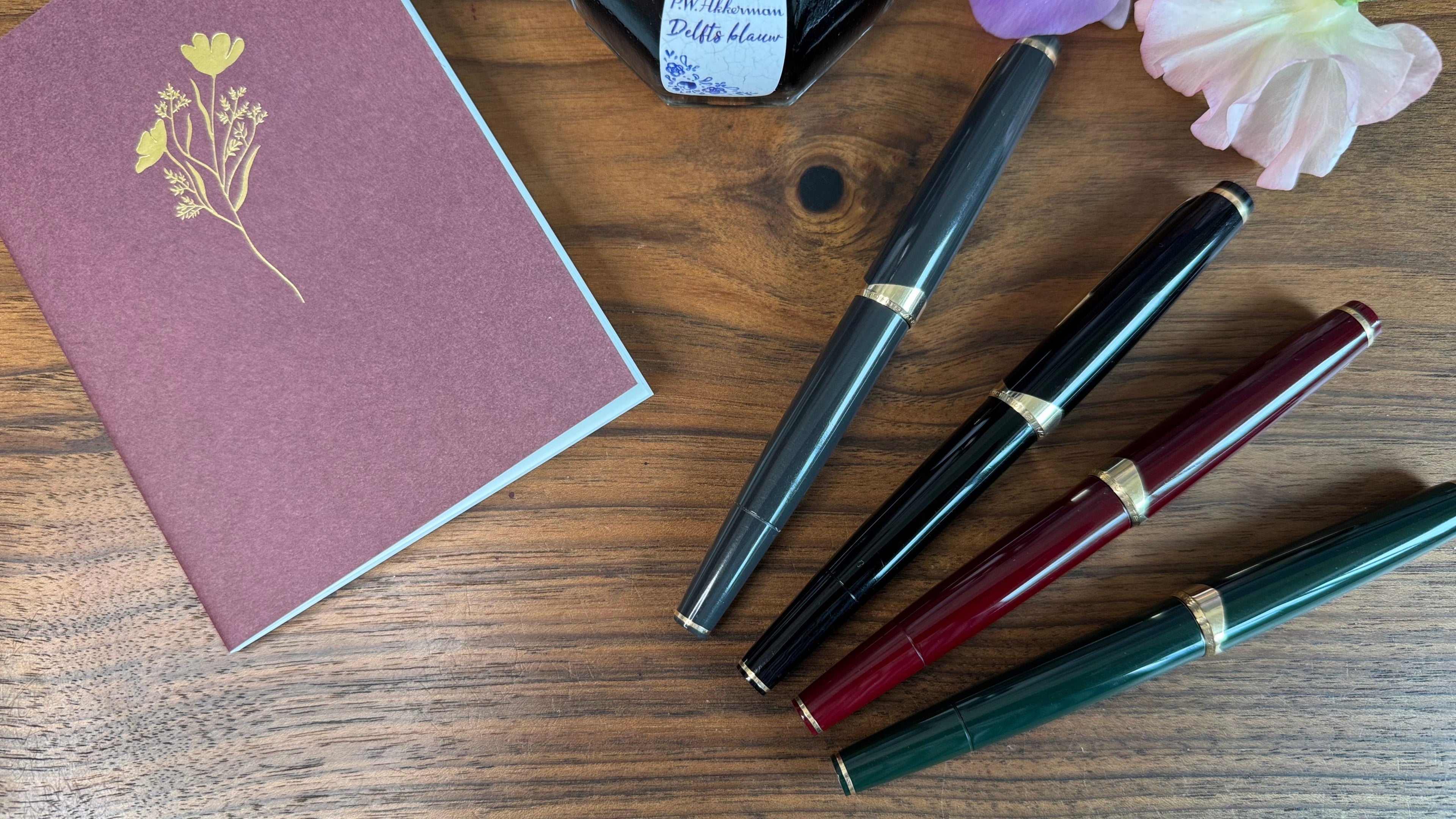 Set of fountain pens and a notebook on a wooden surface with flowers.